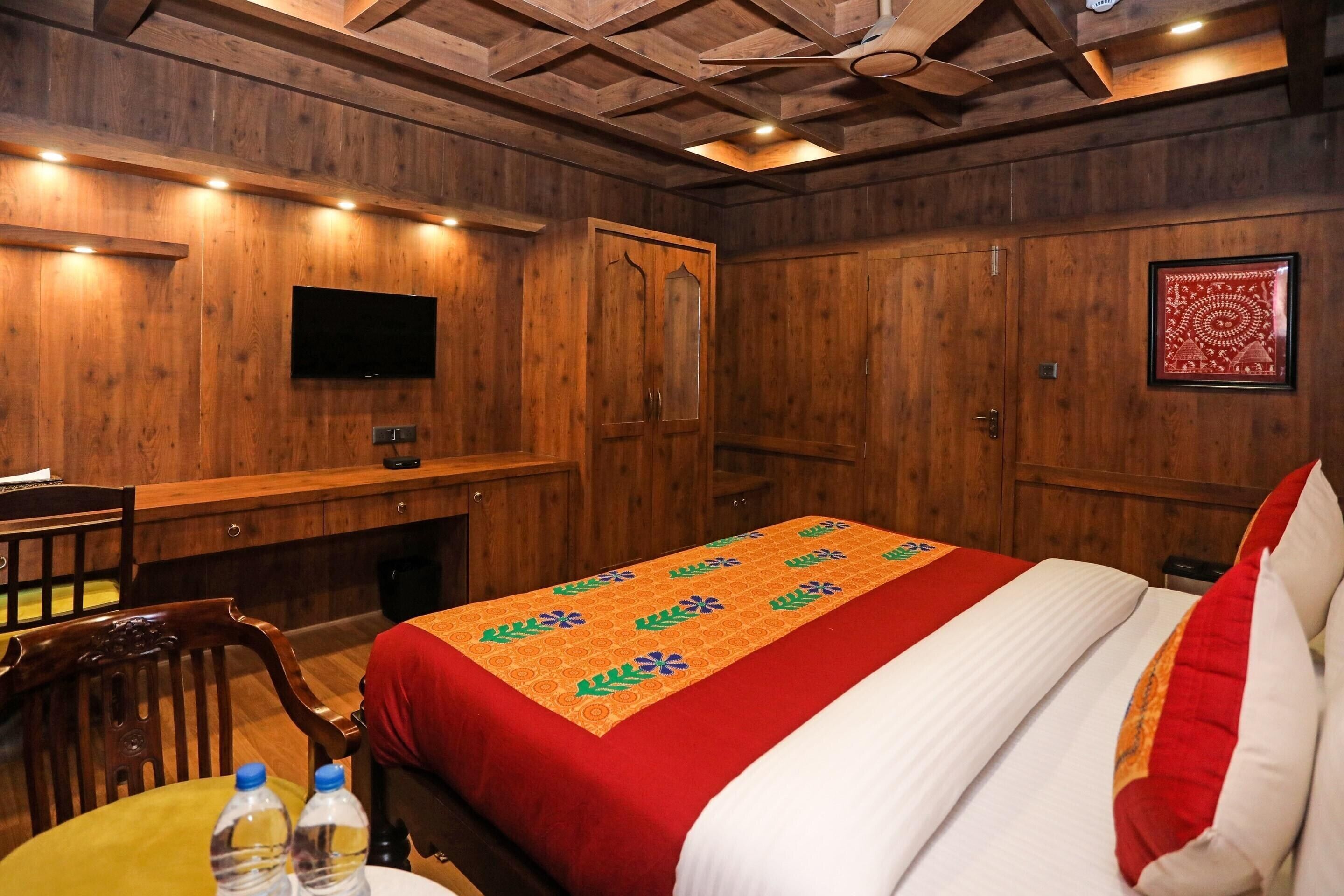 Une chambre d’hôtel avec des boiseries, un lit recouvert d’une courtepointe rouge et blanche, un bureau en bois, une chaise, un téléviseur fixé au mur et une œuvre d’art encadrée.