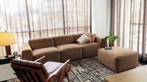 A living room with a brown sofa, a wooden armchair, and a small ottoman. There is a lamp on a side table, a potted plant, and a view through large windows. - w Bright 1BR w View in El Nogal (Bogotá)