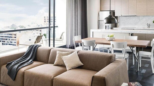 Een moderne woonkamer met een beige bank, een eettafel en stoelen. Er is een keuken met een spoelbak, fornuis en wasmachine. Grote ramen bieden uitzicht op de stad.
