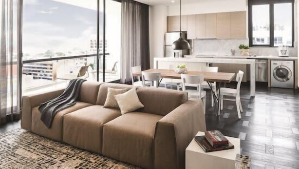 A modern living room with a sofa, dining table, and chairs. A kitchen with a dining area is visible in the background.