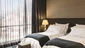 A hotel room with two beds, a large window with sheer curtains, a wooden headboard, and a bedside lamp.