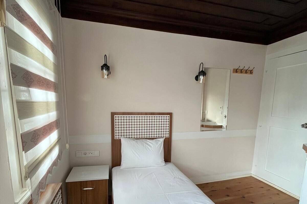 Un dormitorio con cabecera de madera, una mesita de noche, un paralelo, una ventana con persianas, una lámpara de pared y una puerta con perchero.