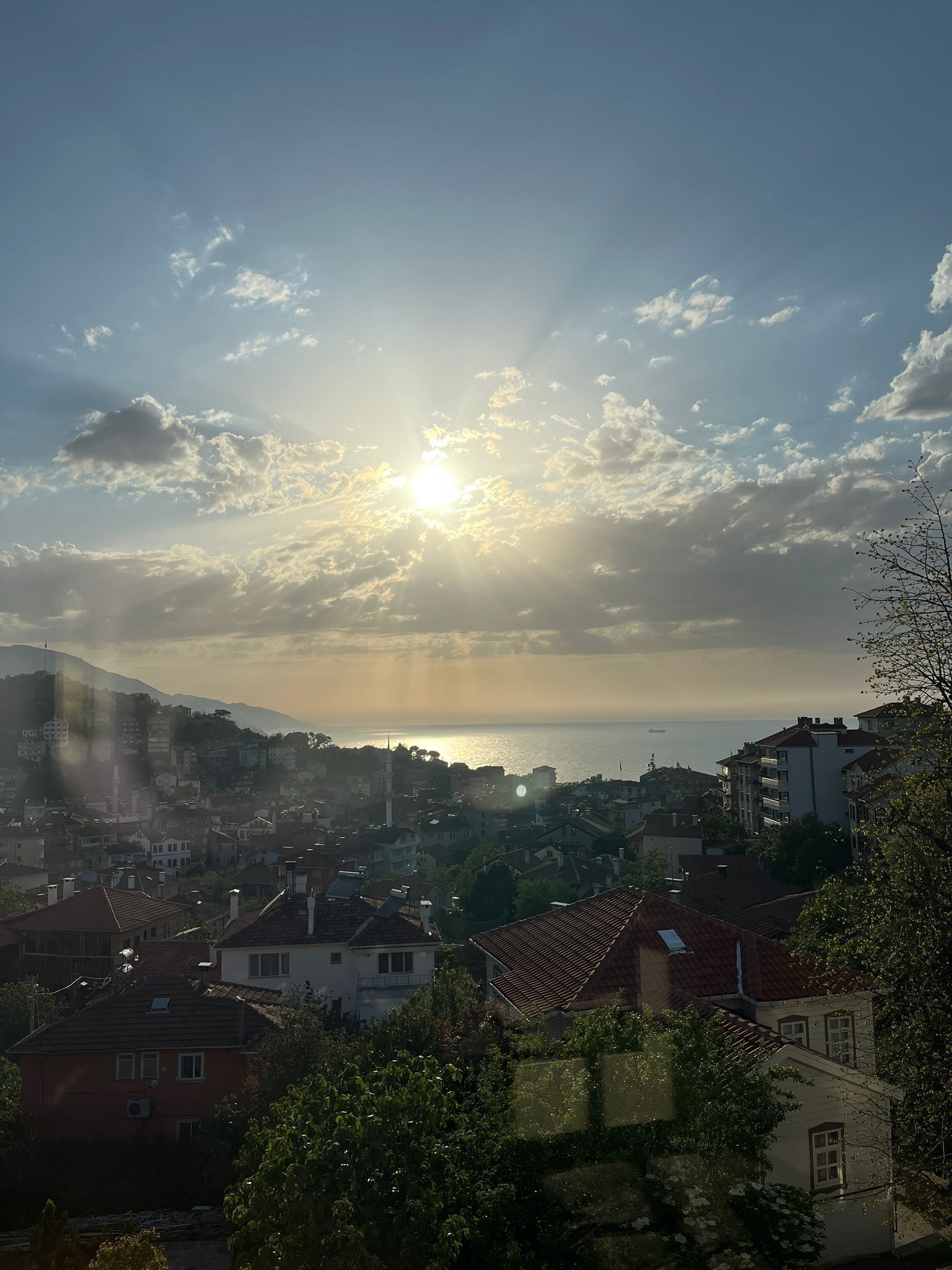 A coastal town with buildings, a mountain in the background, and the sun setting over the sea.