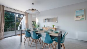 A modern dining room with a large table set for six, surrounded by teal chairs, a hanging light fixture, and a view of the outdoors through large windows.