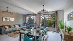 A modern living room with a dining area, large windows, and a view of trees.