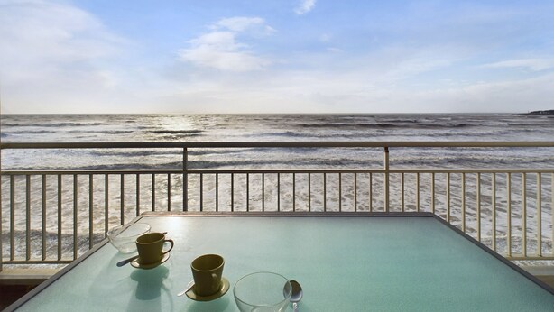 A table with two cups and a spoon, overlooking a beach with waves and a clear sky.