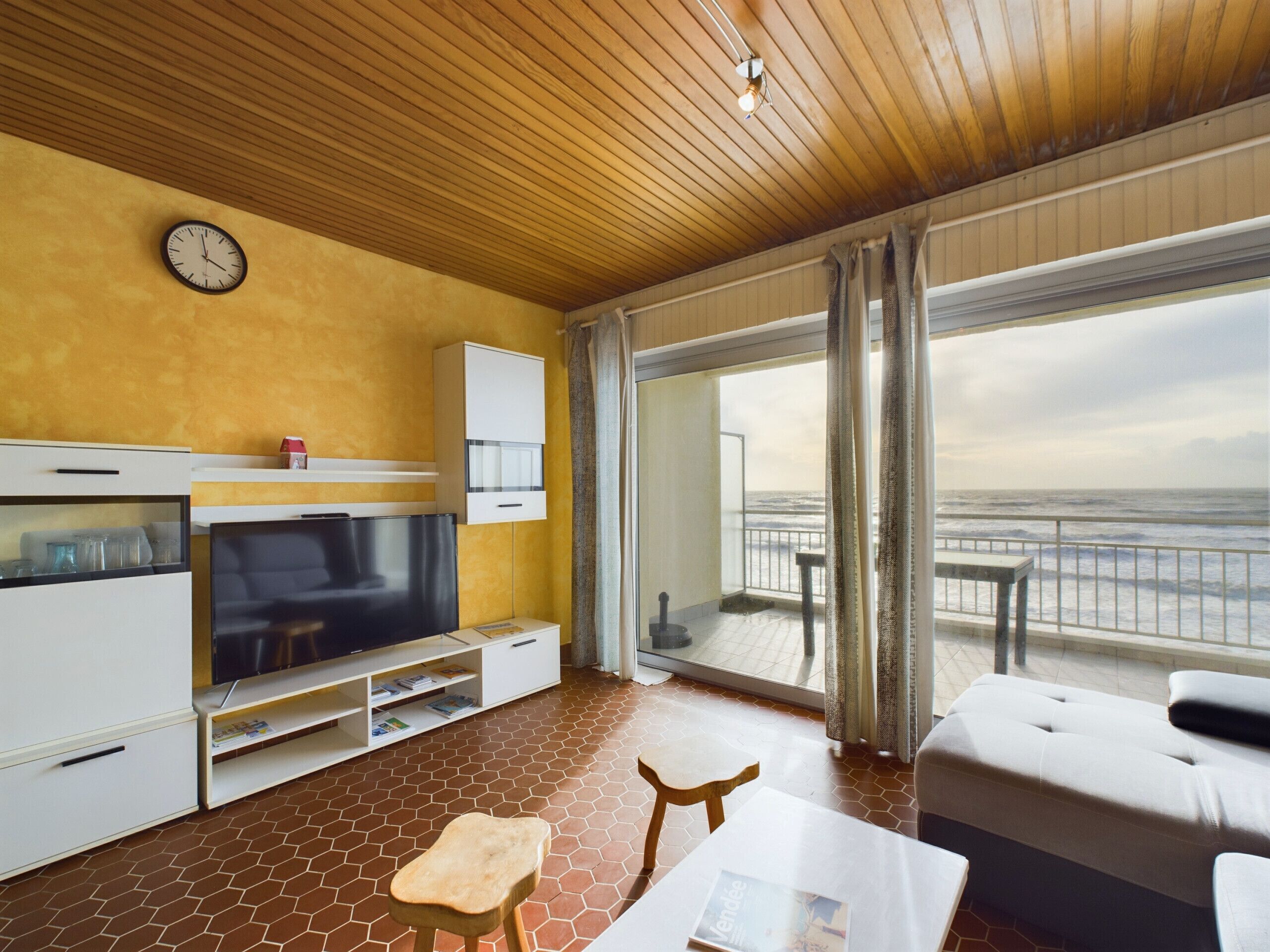 A modern hotel room with a wooden ceiling, a large TV, a sofa, a dining table, and a view of the sea.