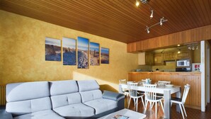 A modern living room with a grey sectional sofa, a white coffee table, and a dining area with white chairs. The kitchen features wooden cabinets and a microwave.