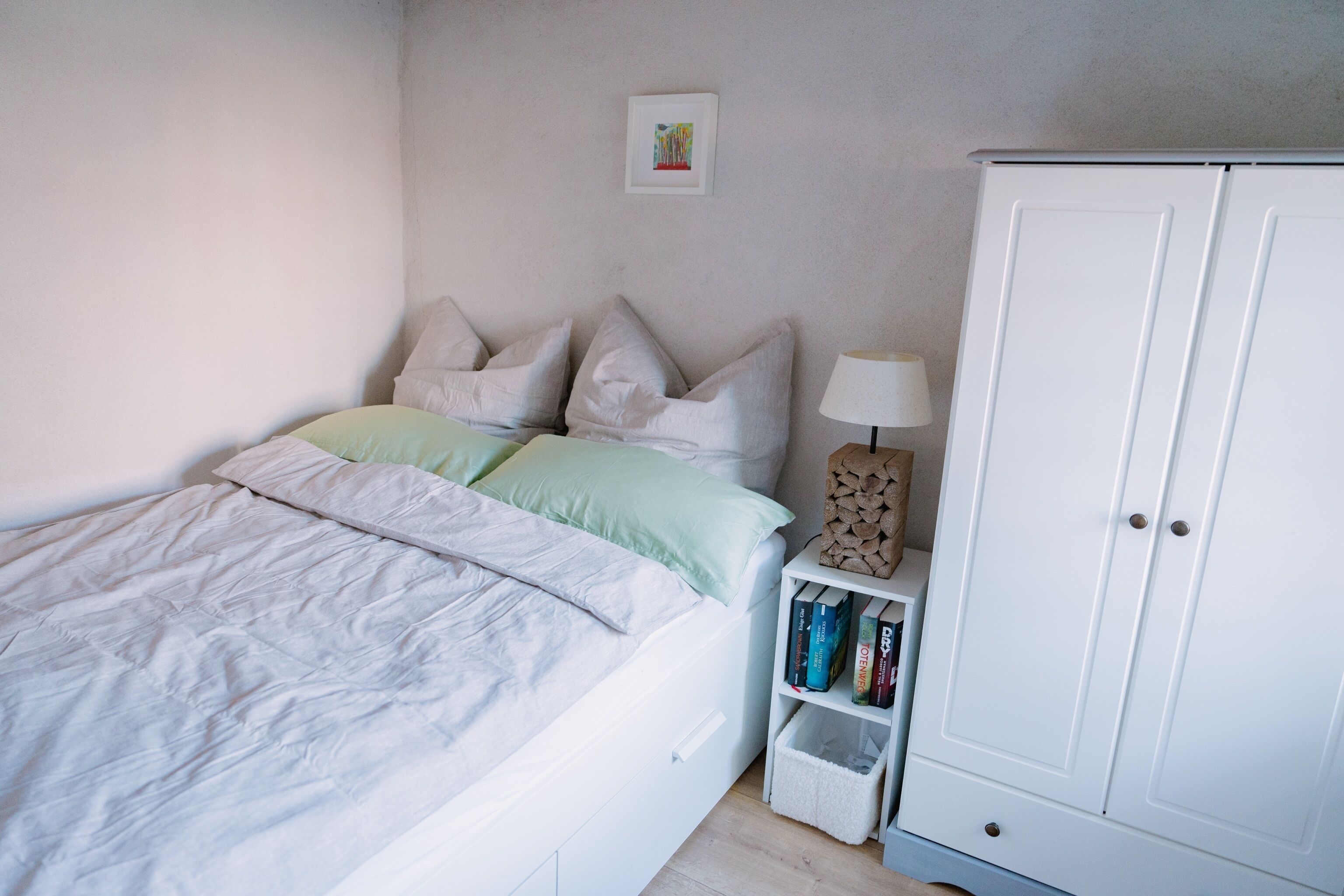 Une chambre à coucher avec un lit, des oreillers, une table de chevet avec des livres, une lampe et une armoire.