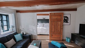 Una sala moderna con un sofá, una mesa de centro y un televisor. Hay una puerta de madera que conduce a un dormitorio con una cama y un ventilador de techo.