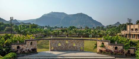 A paved area with a stone wall and arched openings, surrounded by greenery and a mountainous backdrop.