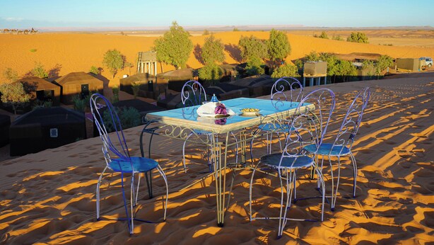 A desert setting with a table and chairs set up for dining, overlooking a vast sandy landscape with sparse vegetation and distant hills.