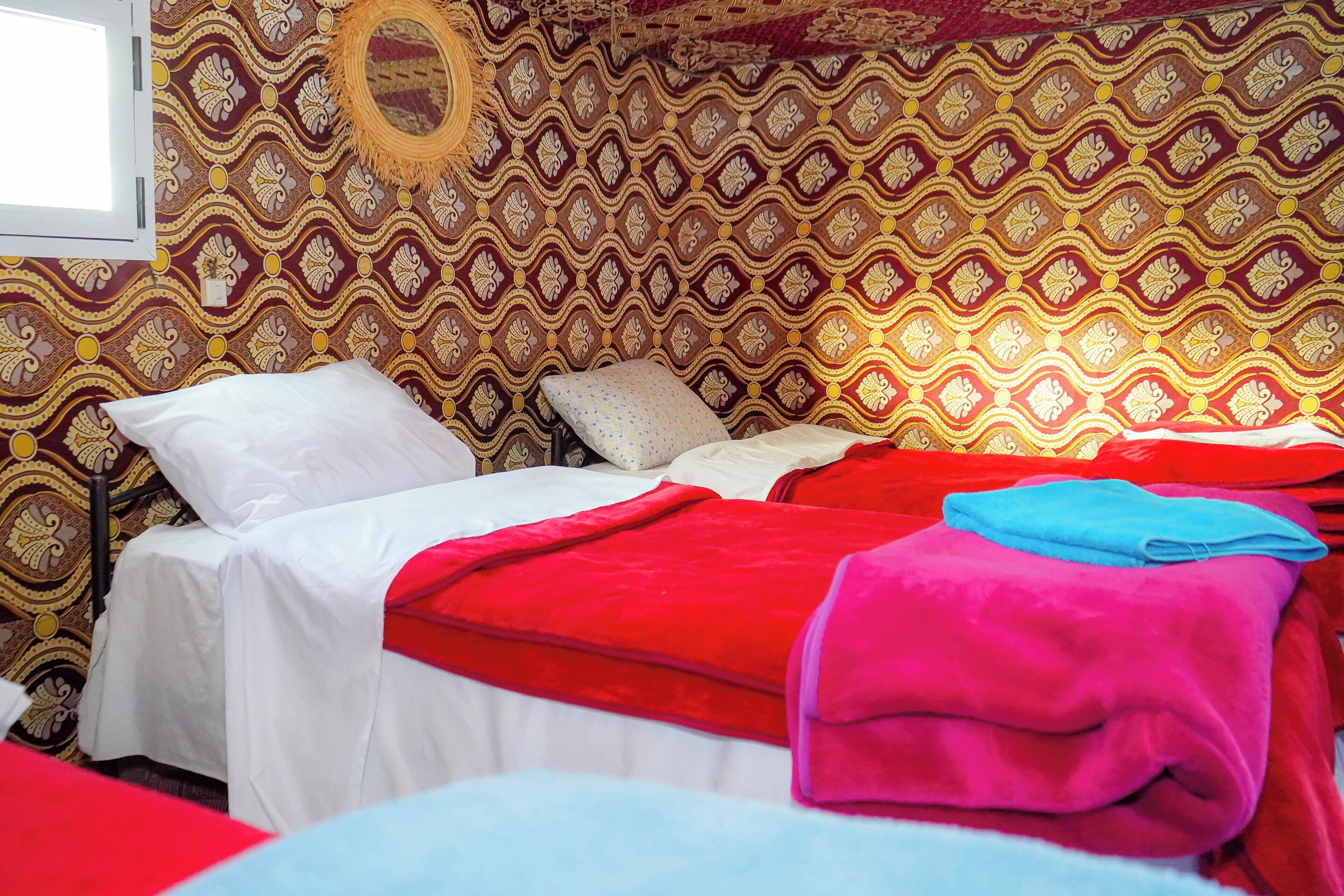Quarto com papel de parede estampado, cama com roupa de cama branca e vermelha, e uma janela.