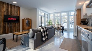 Ein modernes Wohnzimmer mit einer Couch, einer Bank, einem Holzschrank, einem Fernseher, der 'NETFLIX' anzeigt, und einem Essbereich mit Stühlen und einem Tisch.