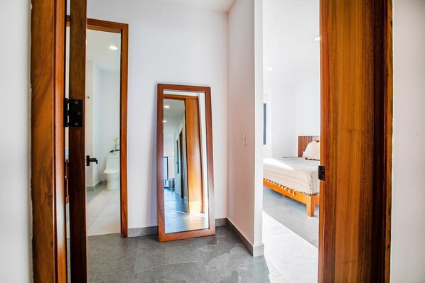 A hallway with a large mirror, a wooden door, and a bed with a wooden frame in the adjacent room.