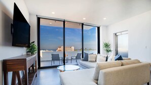A modern living room with a sofa, coffee table, and a TV on a wooden console. Large sliding glass doors lead to an outdoor balcony with a view of the sea and buildings. - Torre Doze Private Penthouse (Puerto Vallarta)