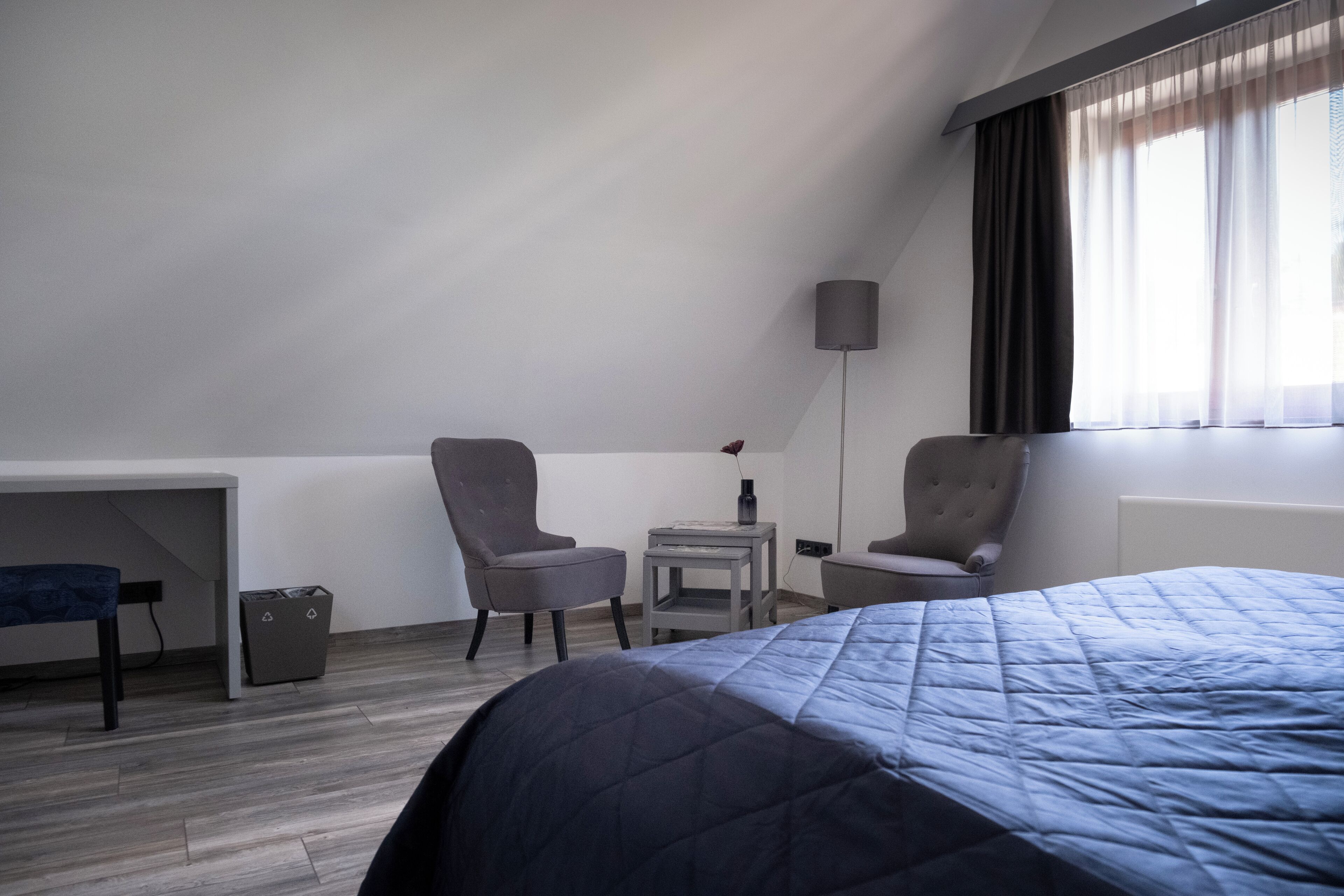 Habitació d'hotel amb un llit, dues butaques, una taula petita, una làmpada, un escriptori i una finestra amb cortines.