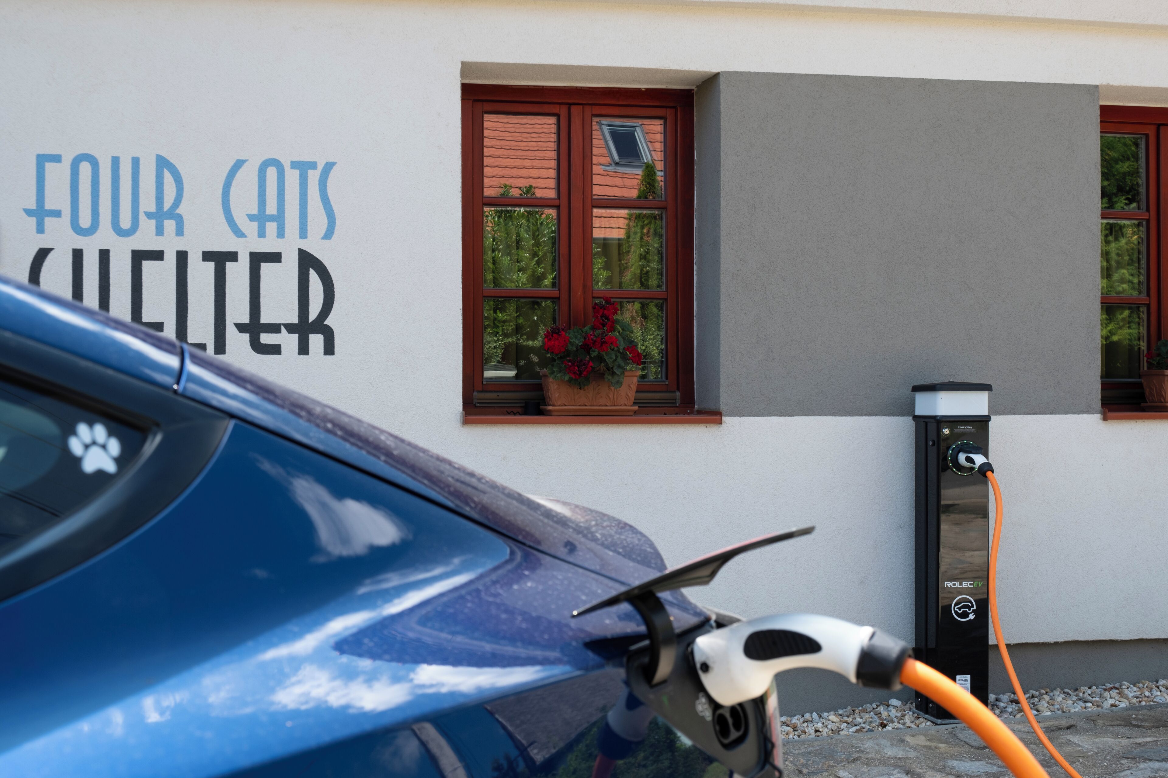 Un coche azul aparcado en una estación de carga frente a un edificio con un cartel que dice "FOUR CATS CLETTER".