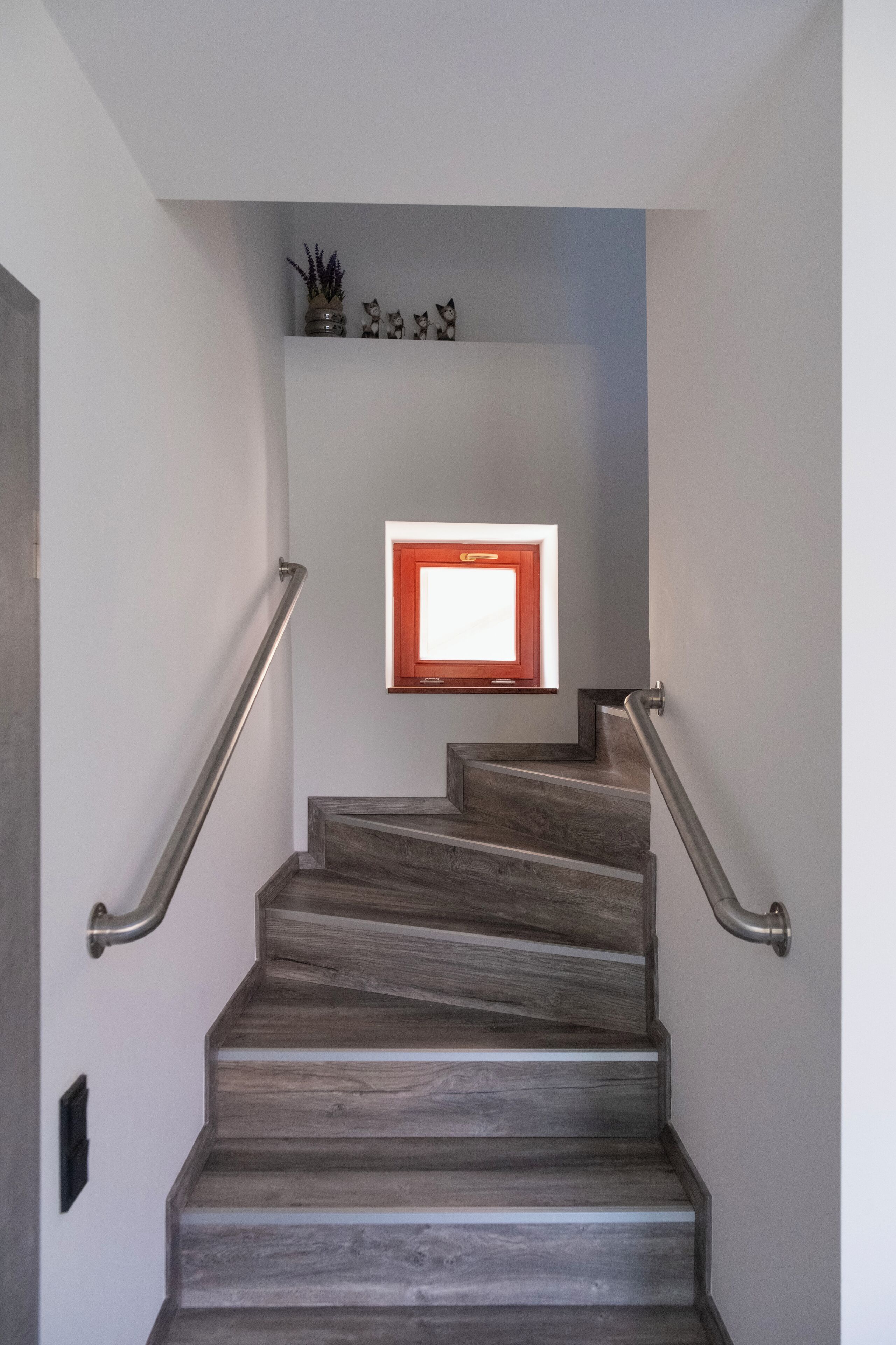 Un escalier avec des marches en bois et des rampes en métal, menant à un palier agrémenté d’une petite fenêtre et d’objets décoratifs.