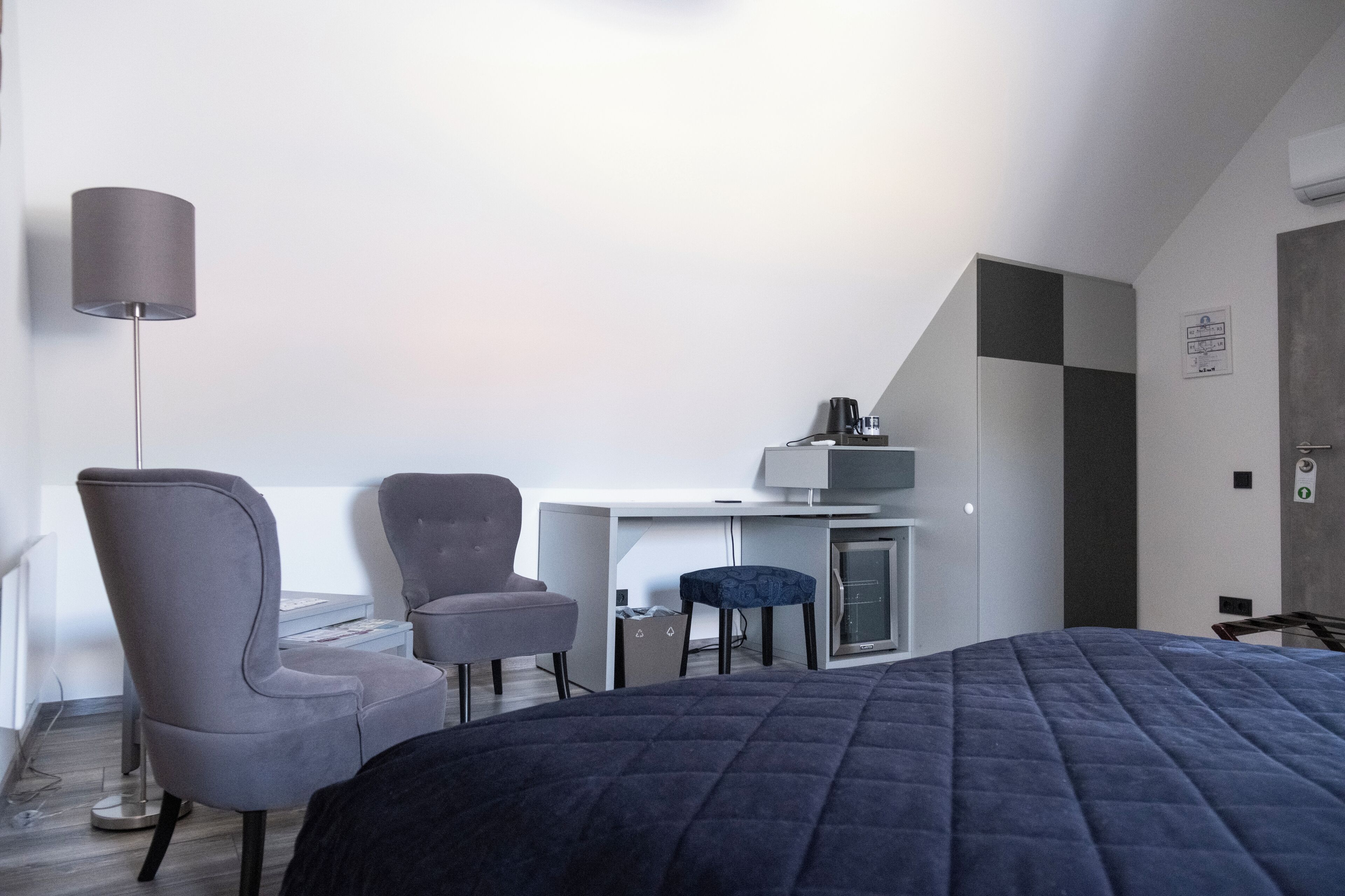Moderni hotellihuone, jossa on sänky, kaksi tuolia, työpöytä tietokoneella, lamppu ja pieni keittokomero.