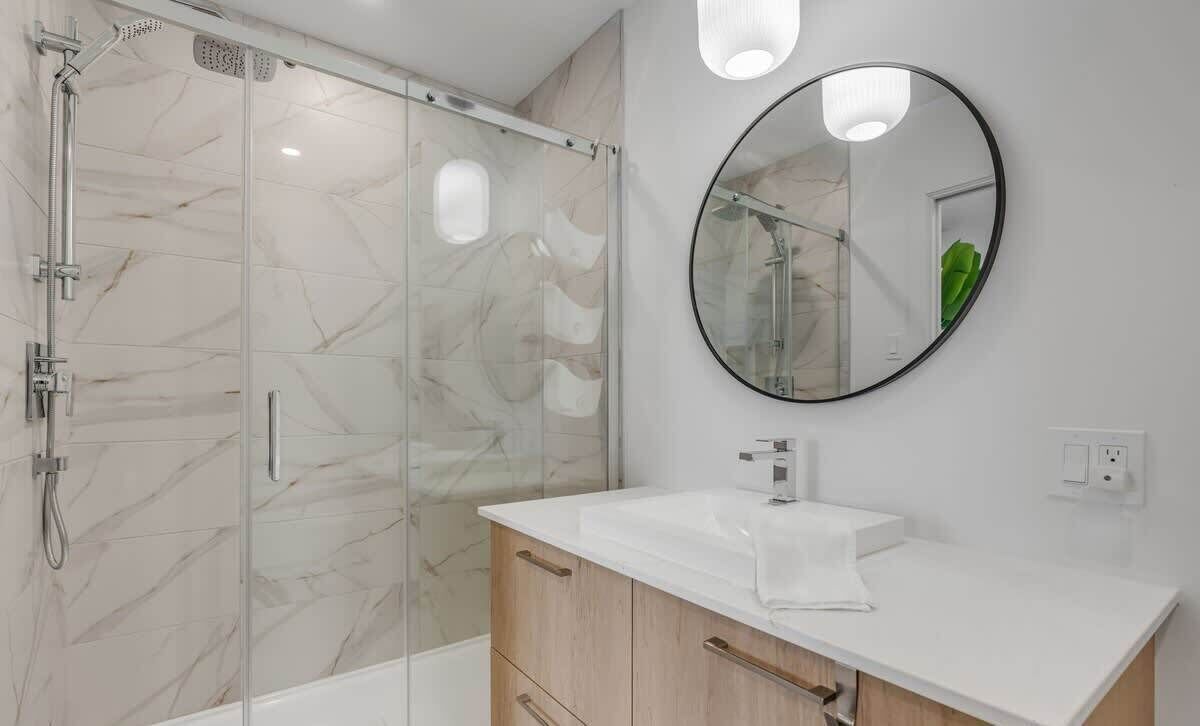 A modern bathroom with a glass-enclosed shower, a white sink, wooden cabinets, and a round mirror.