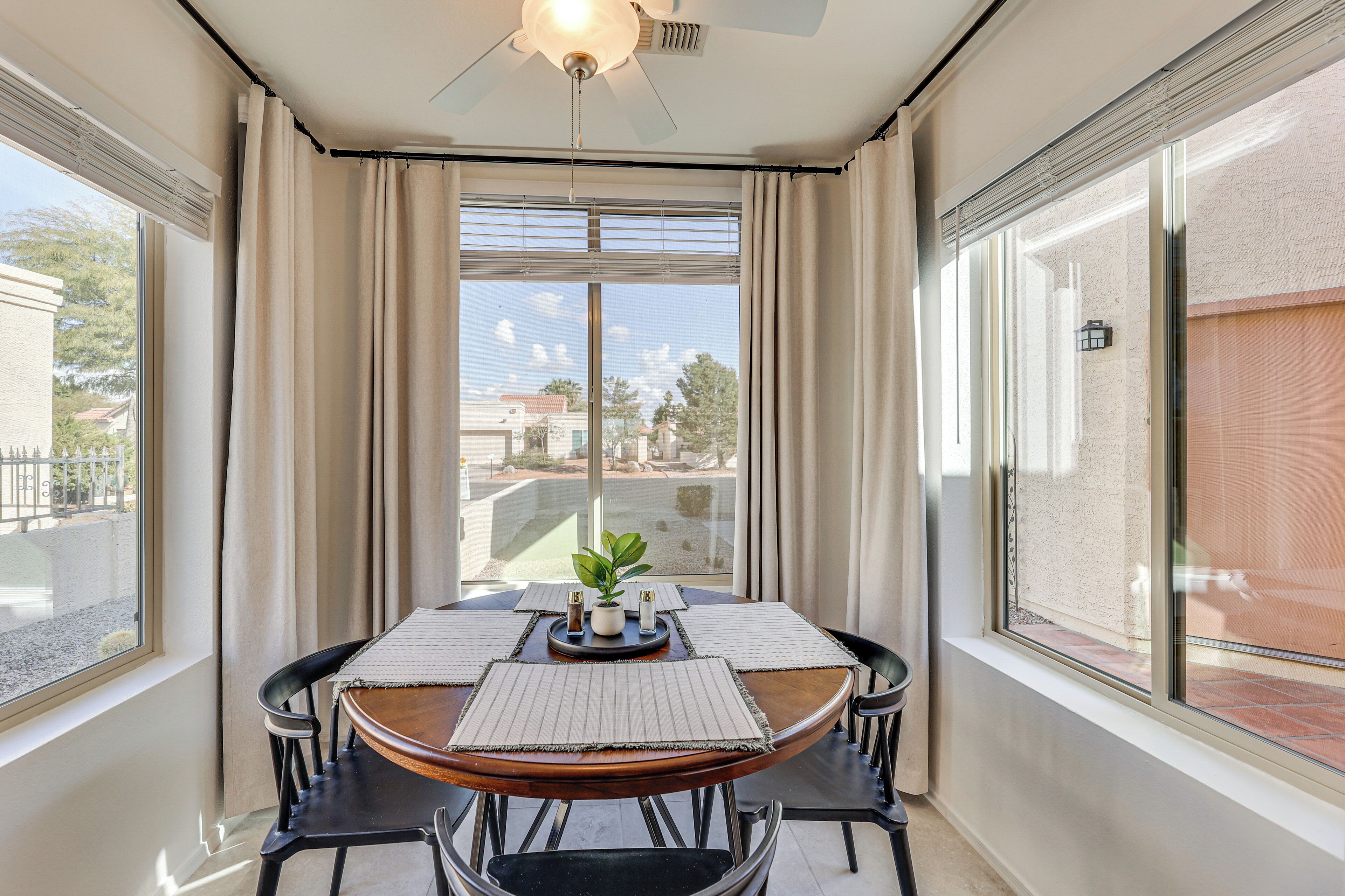 A dining area with a round table, four chairs, a ceiling fan, and a view of buildings through large windows.