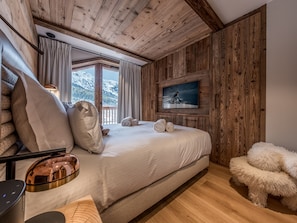 Ein Schlafzimmer mit Holzbalkendecke, einem Bett mit weißen Leinentüchern, einem an der Wand montierten Fernseher und Blick auf schneebedeckte Berge.