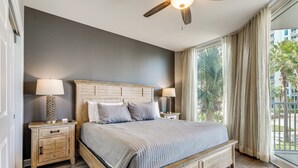 Un dormitorio con una cama de madera, dos mesitas de noche con lámparas, un ventilador de techo y un gran ventanal con cortinas.