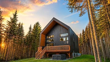 A modern cabin with a wooden deck and large windows, surrounded by a forest.