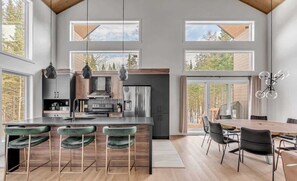 Een moderne keuken met hoge plafonds, grote ramen en een eethoek met een houten tafel en stoelen.