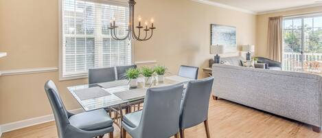 A dining area with a glass-top table, grey upholstered chairs, and a chandelier.