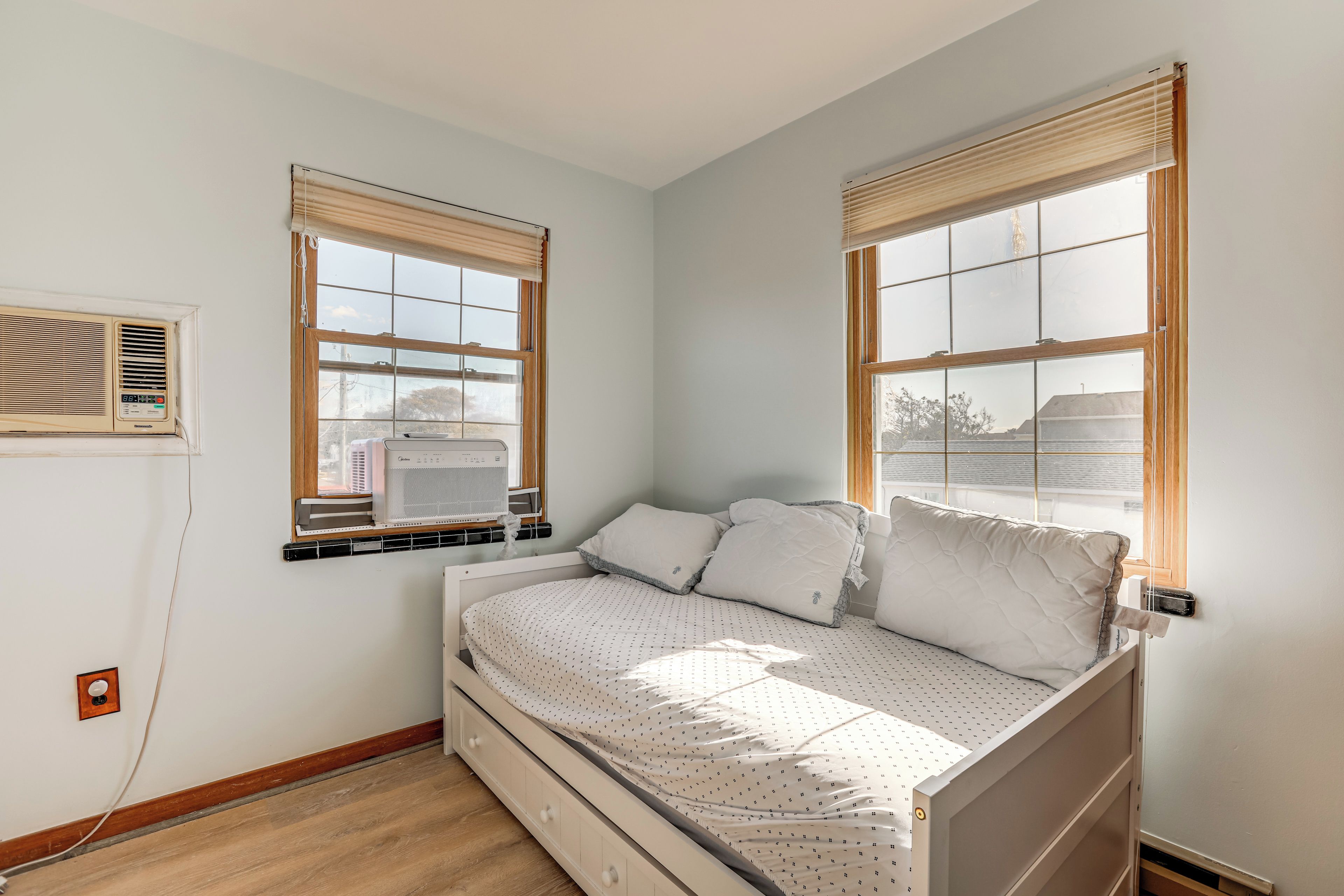 A bedroom with a bed, two windows with blinds, an air conditioning unit, and a wall-mounted heater.