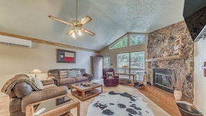 A spacious living room with a leather sofa, a coffee table, and a large cowhide rug. There is a fireplace with a stone wall backdrop and a ceiling fan.