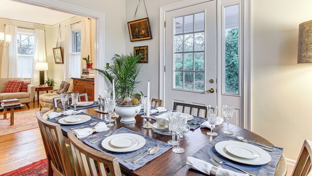 A dining area with a wooden table set for four, surrounded by chairs. There is a potted plant on the table, and the room has a cozy, homey feel with a red rug and a sofa in the background. - 3 Mi to Augusta National: Historic Home w/ Patio (Augusta)