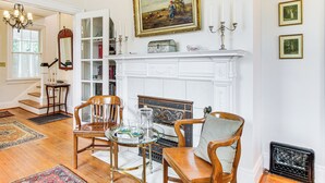 A room with a fireplace, a dining table with chairs, a bookshelf, and a chandelier. - 3 Mi to Augusta National: Historic Home w/ Patio (Augusta)