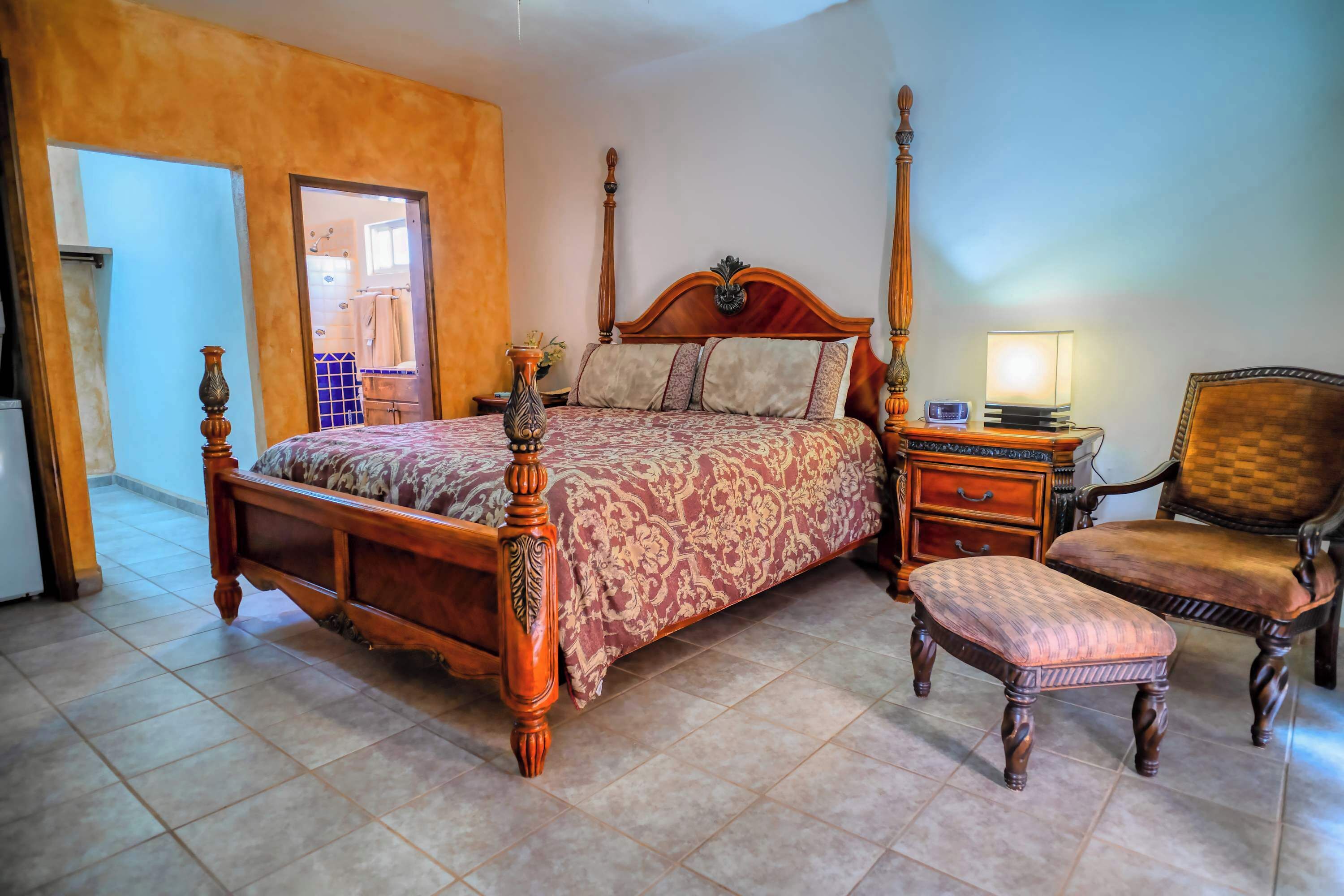 A four-poster bed with a wooden headboard, a nightstand with a lamp, a wooden chair, and a bench.