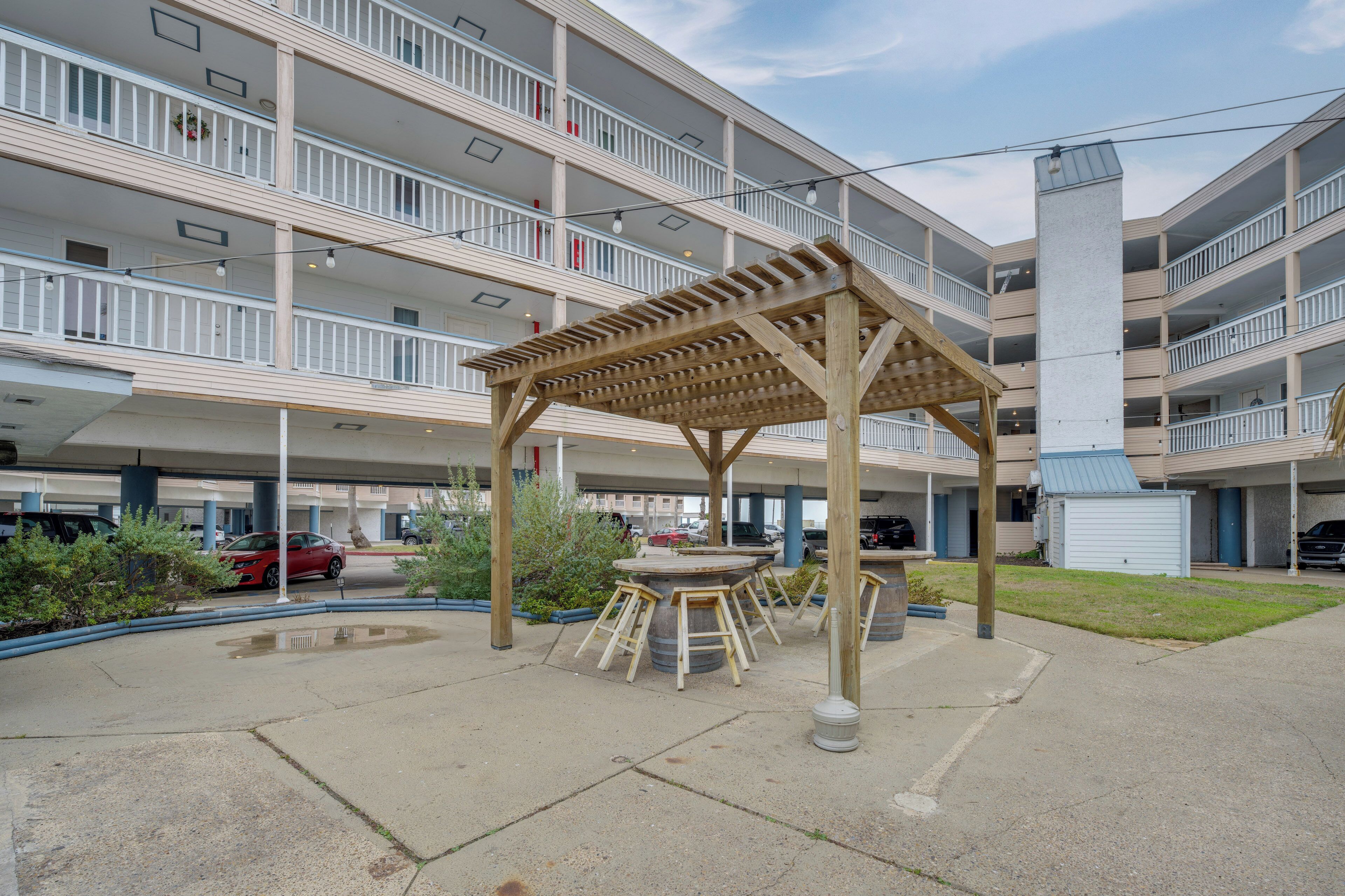 A multi-story building with a covered outdoor seating area, a central column, and a parking lot with cars.