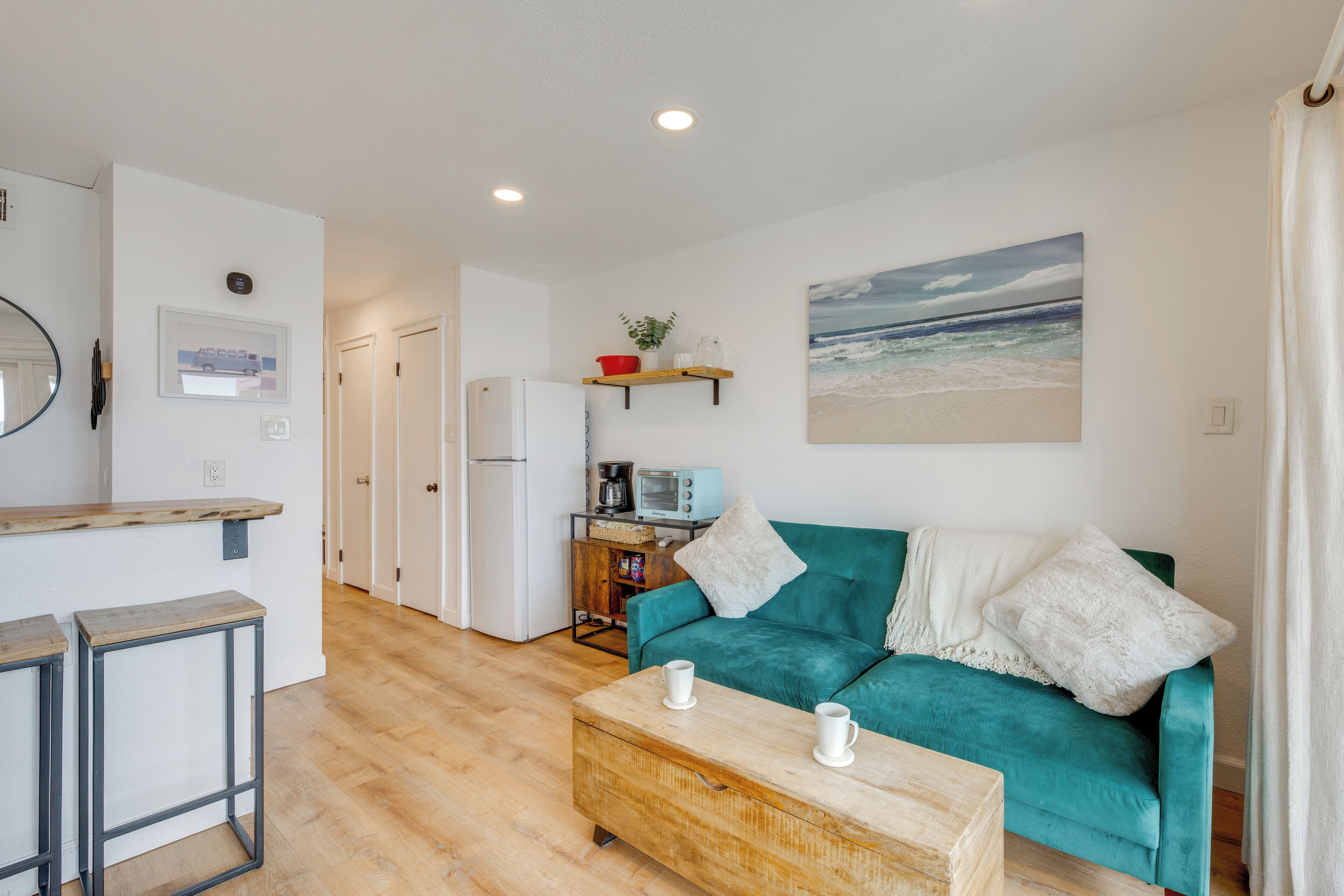 Een ruimtelijk ingerichte leefruimte met kitchenette, een blauwgroene bank, een houten bijzettafeltje en een decoratie met strandthema.