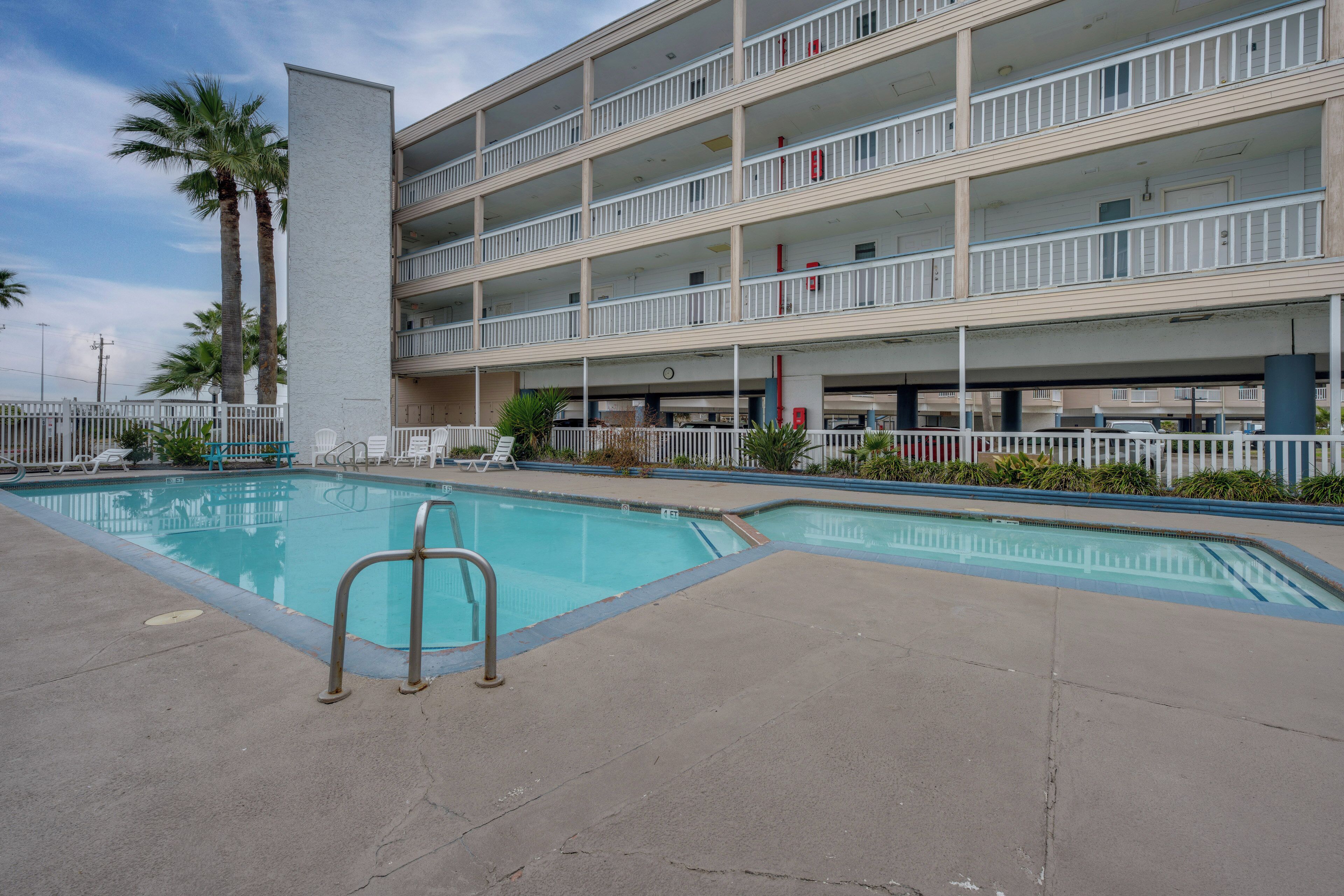 A multi-story hotel with a swimming pool, palm trees, and a white fence.