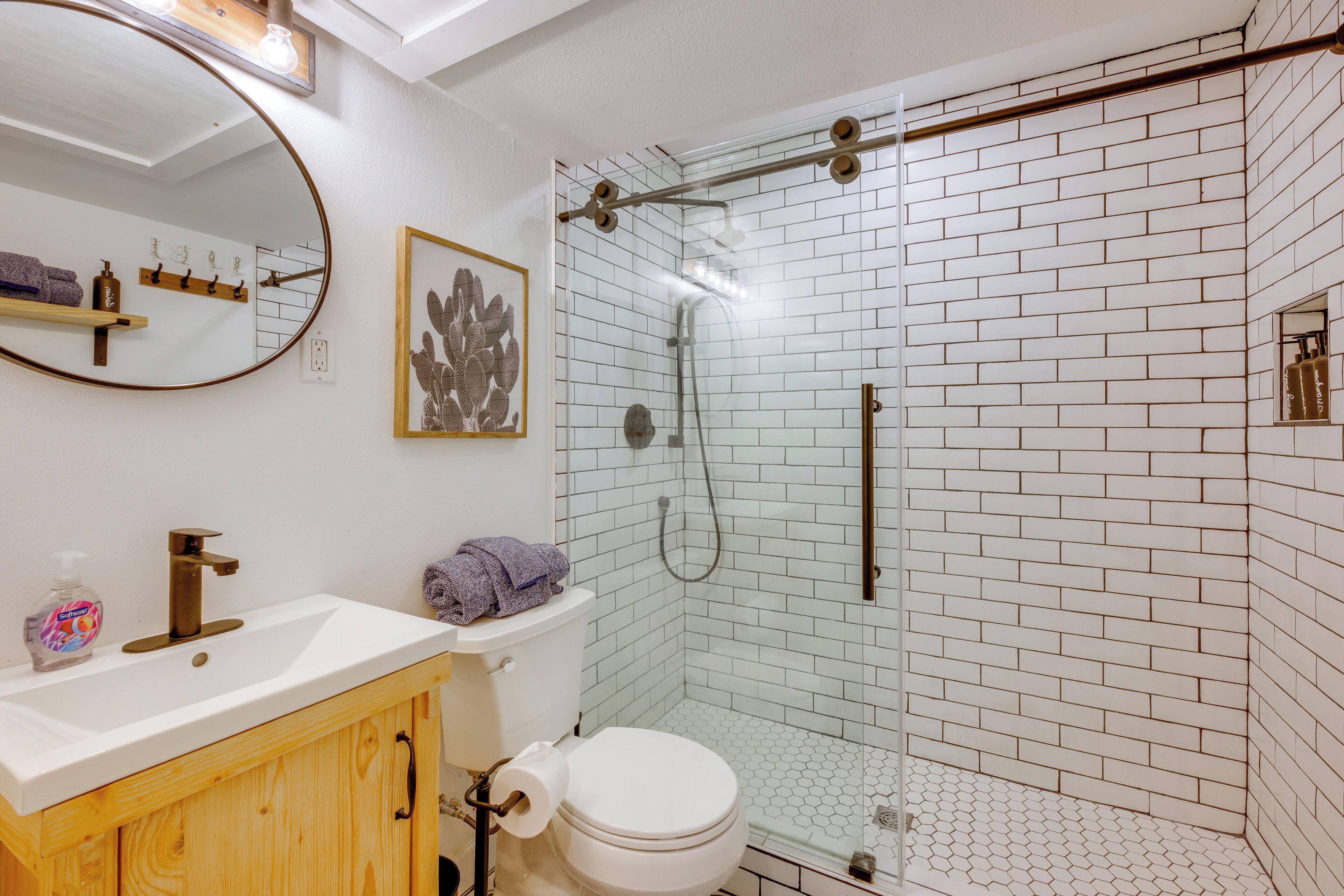 A bathroom with a white sink, wooden cabinet, toilet, and a glass shower enclosure with white tiled walls.