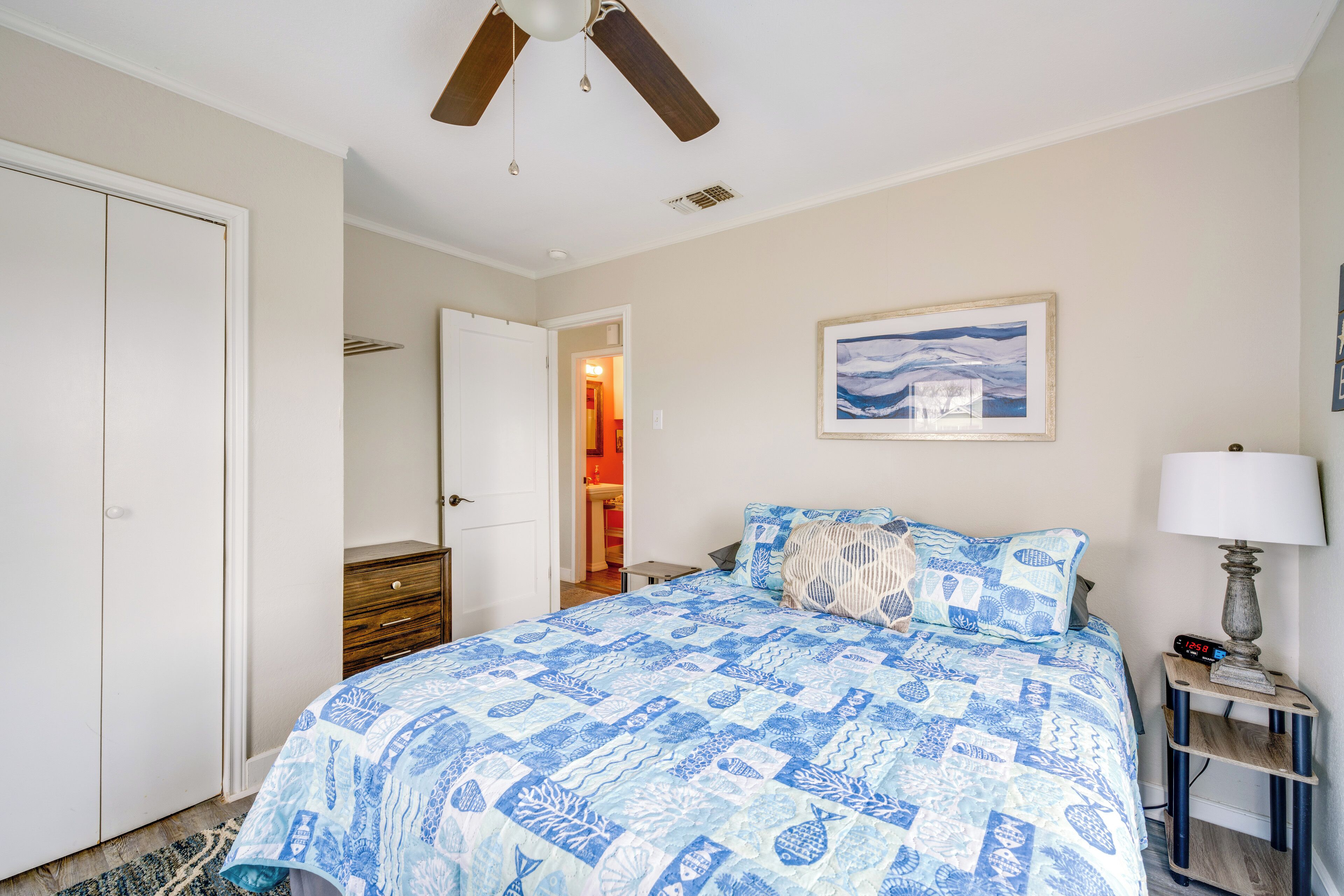 A bedroom with a bed, a ceiling fan, a nightstand with a lamp, a framed picture on the wall, and a door leading to another room.