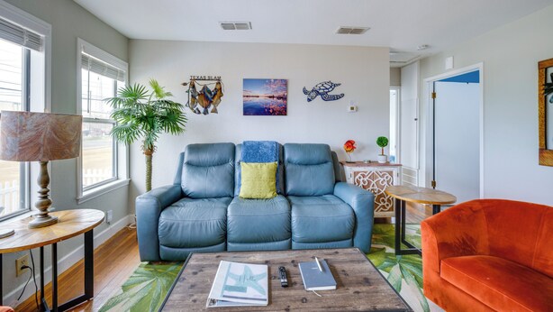 Una sala de estar con un sofá azul, un sillón naranja, una mesa de centro con libros y un control remoto, una lámpara y una planta en maceta.