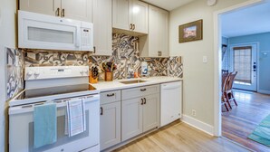 Una cocina moderna con armarios blancos, microondas, estufa y lavavajillas. Tiene un salpicadero decorativo y una ventana con persianas.