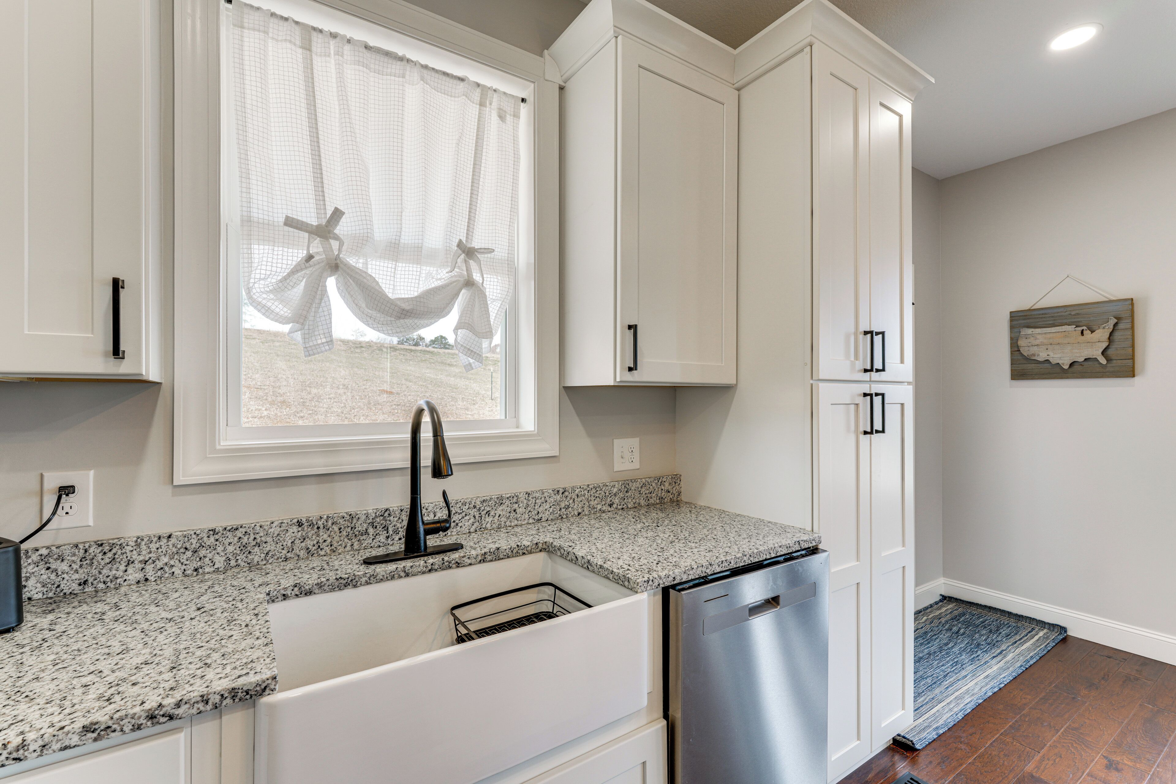 A kitchen with white cabinets, a granite countertop, a stainless steel dishwasher, and a window with a view of a hammock.