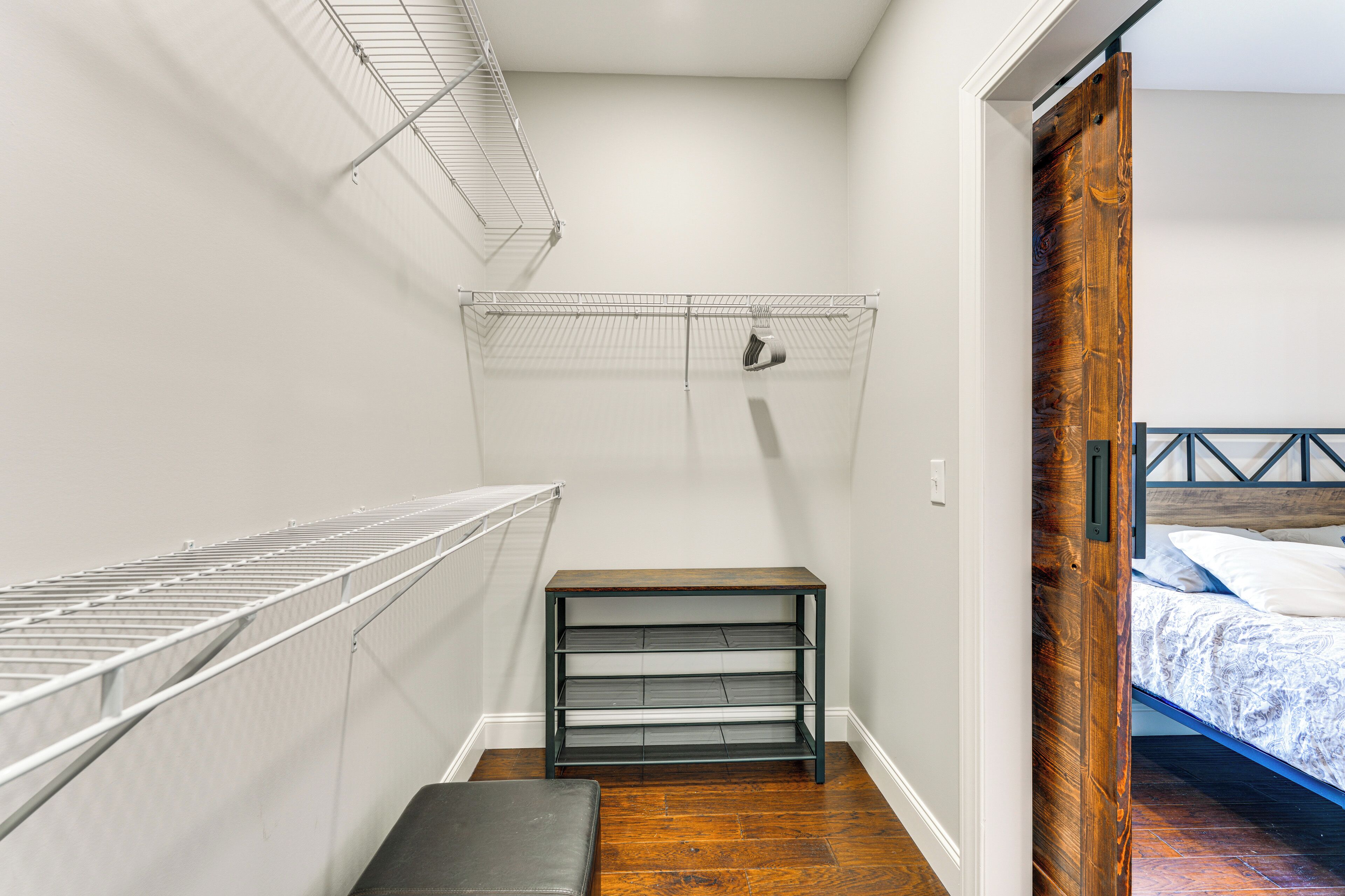 A small, narrow room with a wooden bench, a metal shelf unit, and a bed with a wooden headboard.