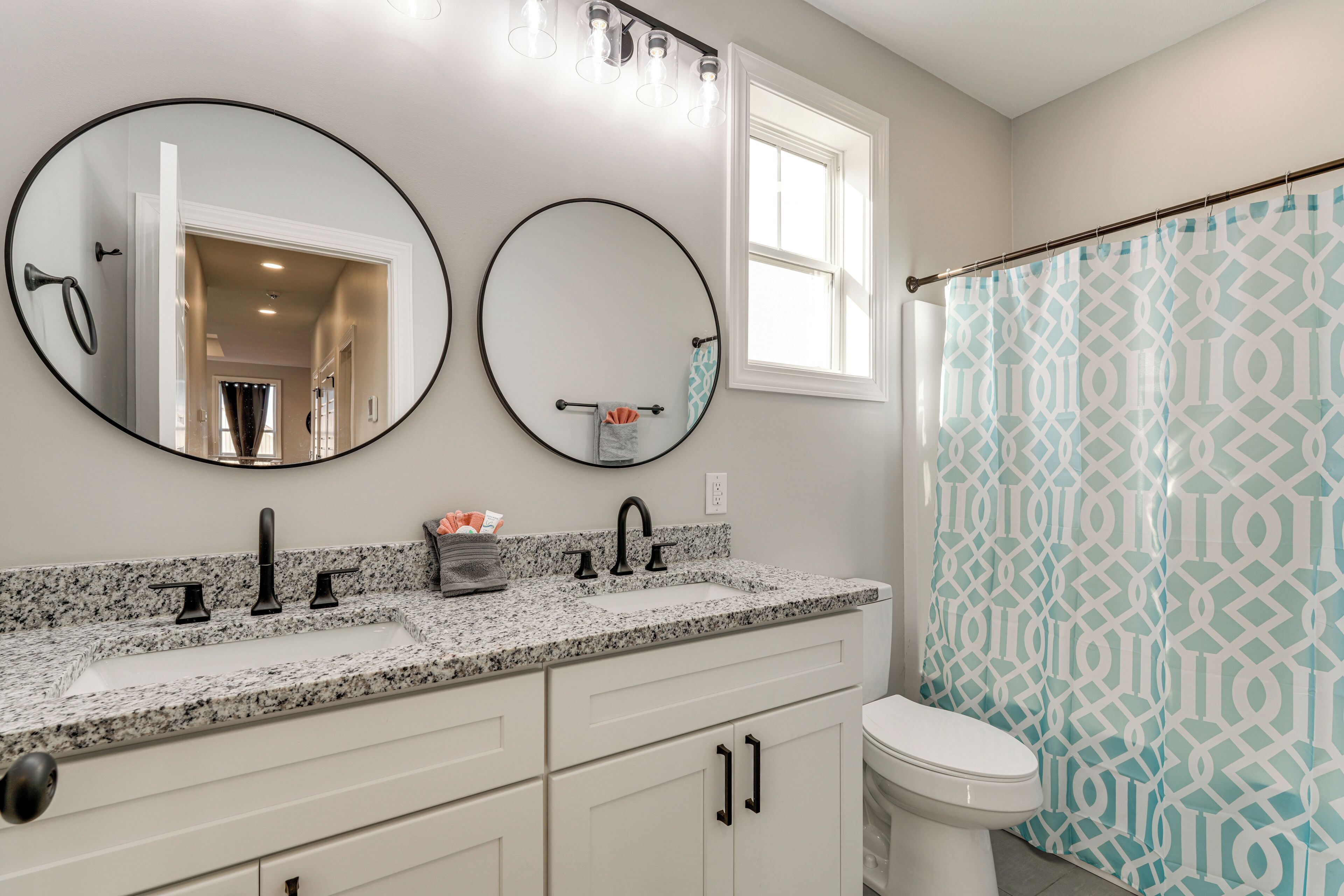 A bathroom with two round mirrors, a white sink with a granite countertop, a toilet, and a shower curtain with a geometric pattern.