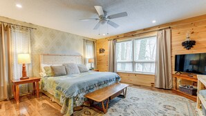 Un dormitorio con cama, piso de madera, ventilador de techo, ventana con cortinas y un televisor sobre un mueble de madera.