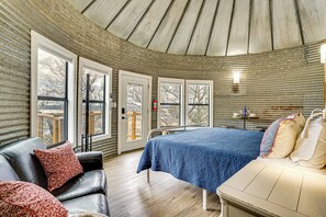 Ein Zimmer mit gewölbter Decke, einem Bett mit blauer Steudecke, einem Ledersofa mit roten Kissen und großen Fenstern.