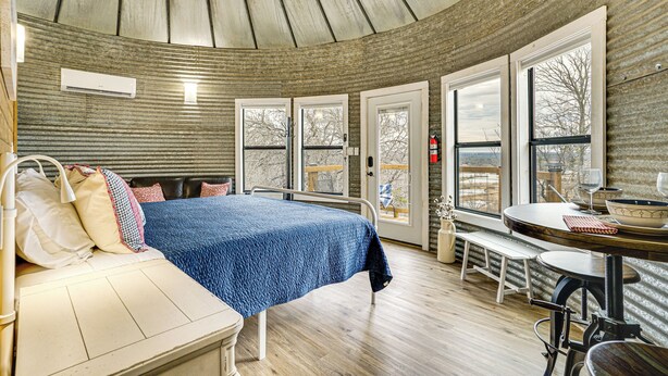 A cozy room with a bed, a bench, a table with a bowl and wine glasses, and large windows offering a view. - Tin De Blue Grainbury Silo and Cabins (Weatherford)