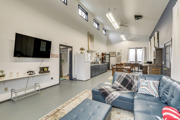A spacious living room with a blue leather sofa, a flat-screen TV mounted on the wall, and a kitchen area in the background. - 5 Mi to Hillsboro: Vintage-style Home w/ Fire Pit (Cornelius)